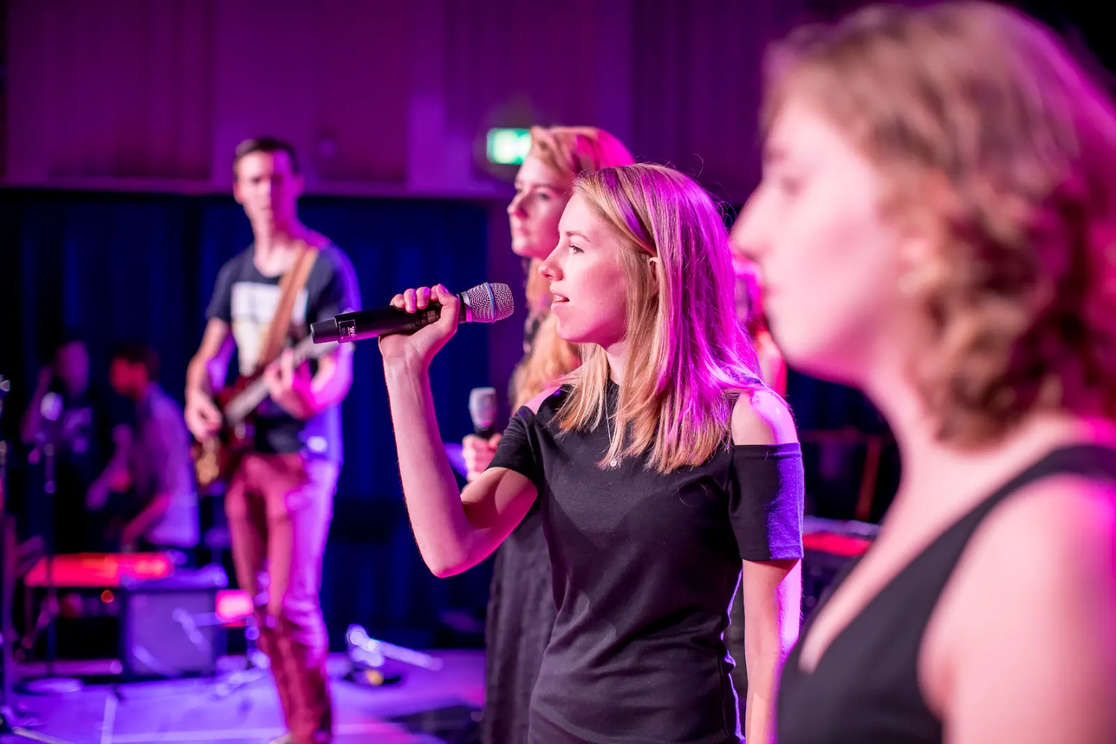 Zangeressen vooraan op het podium, gitarist erachter, paars licht